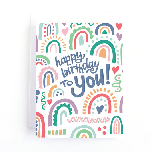 Birthday card for her with the hand lettered text, Happy Birthday to you! surrounded by illustrations of whimisical rainbows.