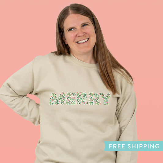 Woman wearing a beige sweatshirt with holiday greenery forming the word 'MERRY'