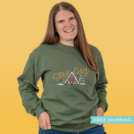 Woman wearing a green sweatshirt with 'Stay Cosy' text and snowy winter cabin  in the woods.