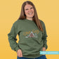Woman wearing a green sweatshirt with 'Stay Cosy' text and snowy winter cabin  in the woods.