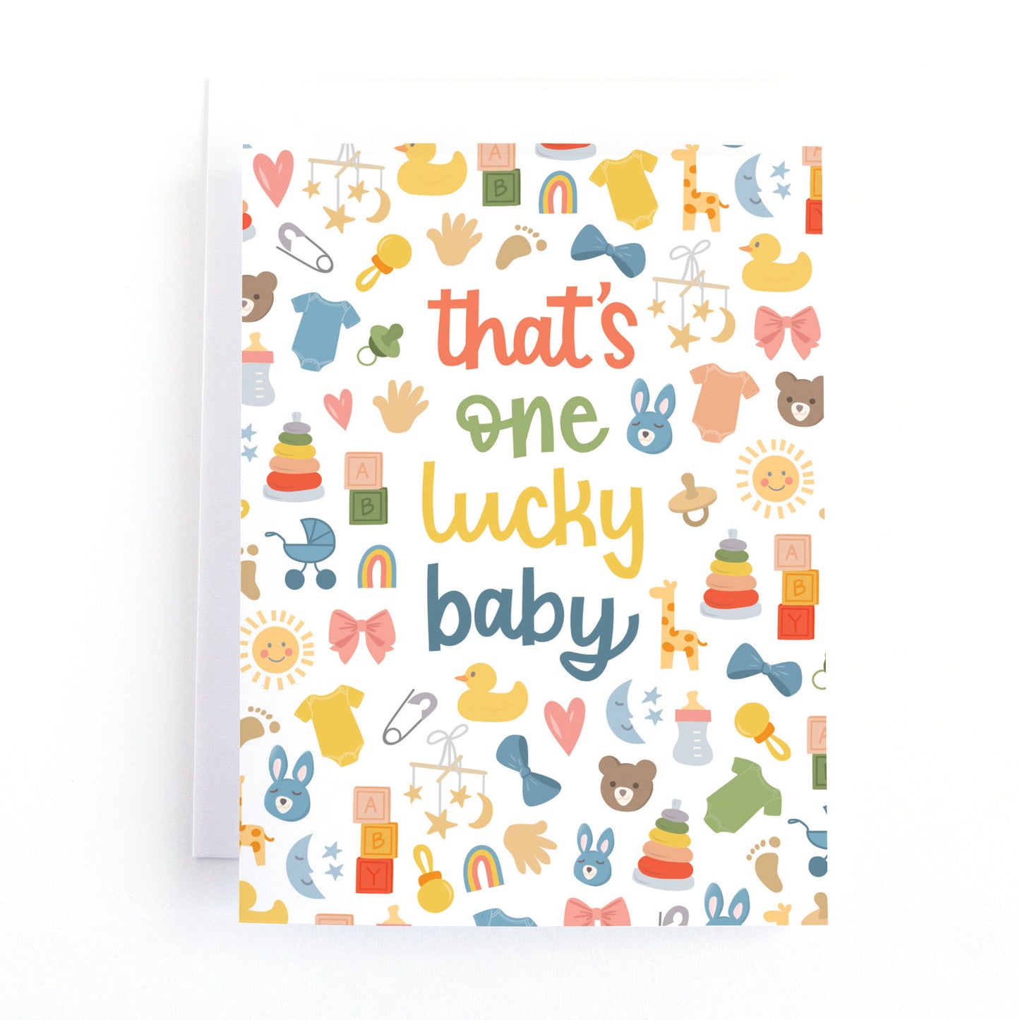 A baby shower card with the phrase 'that's one lucky baby' in colorful text, surrounded by a playful pattern of baby-themed illustrations including ducks, bows, and toys, shown with a white envelope on a white background.