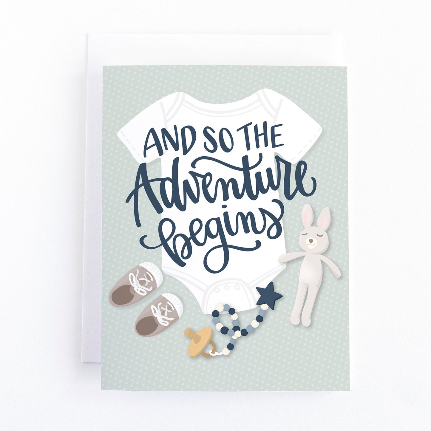 Gender neutral baby shower card with a flat lay of baby layette including a onsie, baby shoes, pacifer and soft toy with the message "and so the adventure begins"