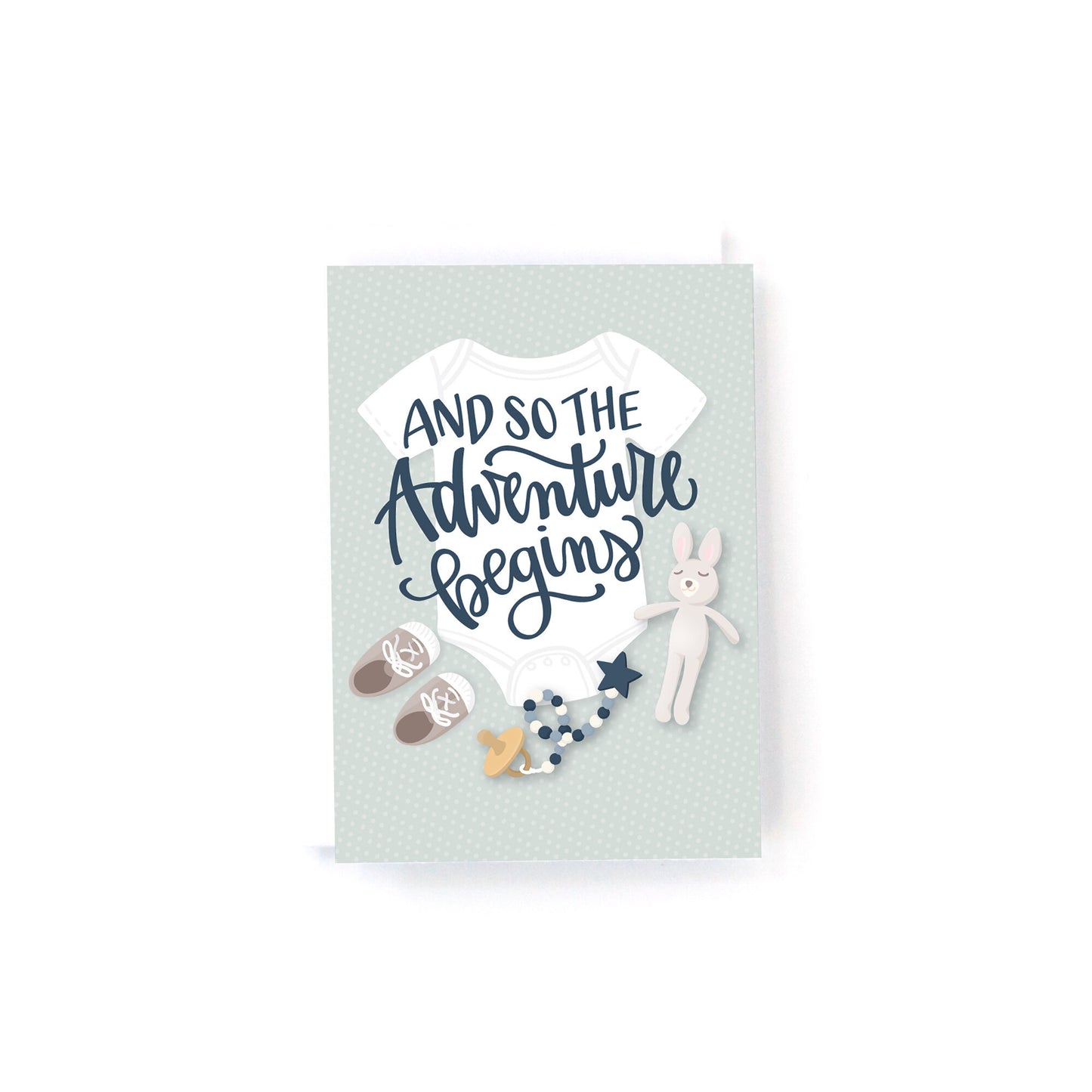 mini new baby card with illustrations of a baby layette on a neutral polka dot background and the text, and so the adventure begins.