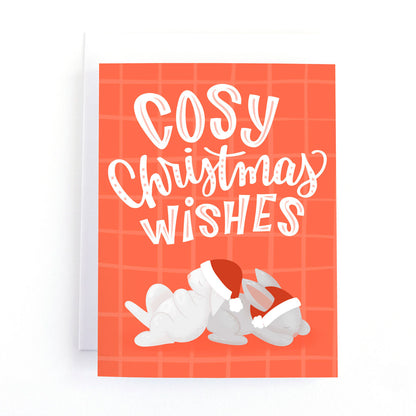 bunny christmas card featuring two rabbits wearing santa hats all cuddled up for a nap with the hand lettered message, cosy christmas wishes.